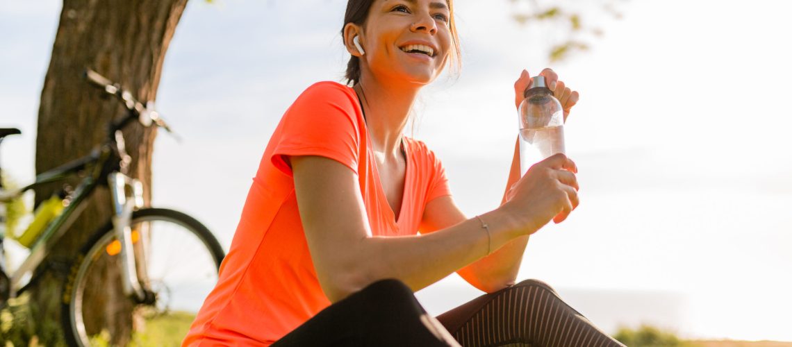smiling beautiful woman drinking water in bottle doing sports in morning in park nature doing yoga on mat, pink fitness outfit, happy healthy lifestyle, music in earphones, bicycle on background